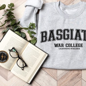 May include: A grey sweatshirt with the text "BASGIATH WAR COLLEGE LIGHTNING WIELDER" in black. An open book, glasses, and a patch with the words "LIGHTNING WIELDER" are also visible.
