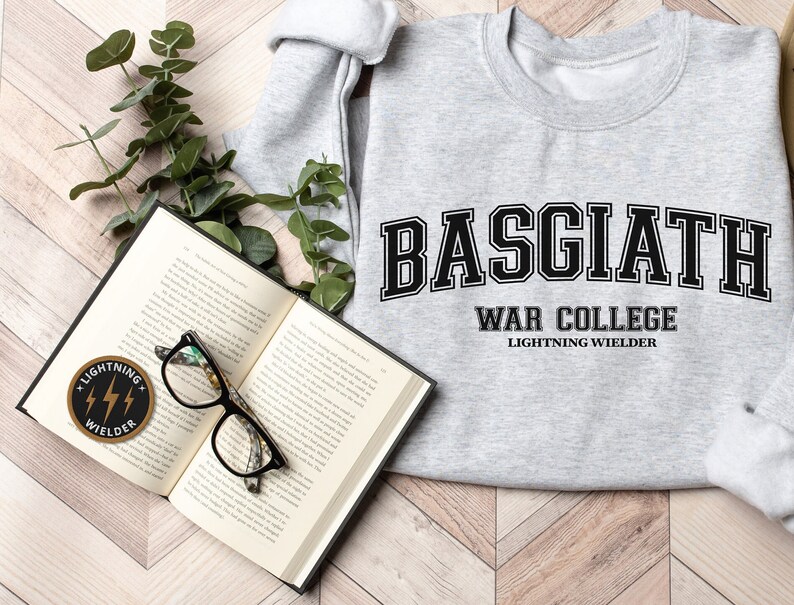 May include: A grey sweatshirt with the text "BASGIATH WAR COLLEGE LIGHTNING WIELDER" in black. An open book, glasses, and a patch with the words "LIGHTNING WIELDER" are also visible.