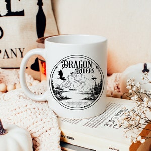 May include: White ceramic mug with a black and white graphic of a dragon flying over a mountain range. The graphic is surrounded by a circle with the text "Dragon Riders" in a black font. The text below the graphic reads "A dragon without its rider is a tragedy. A rider without its dragon is dead."