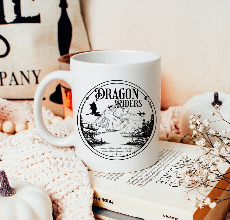 May include: White ceramic mug with a black and white graphic of a dragon flying over a mountain range. The graphic is surrounded by a circle with the text "Dragon Riders" in a black font. The text below the graphic reads "A dragon without its rider is a tragedy. A rider without its dragon is dead."