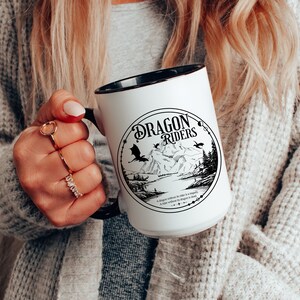 May include: White ceramic mug with a black rim and handle. The mug features a black and white illustration of a mountain scene with two dragons flying overhead. The text "Dragon Riders" is printed in a black, stylized font.