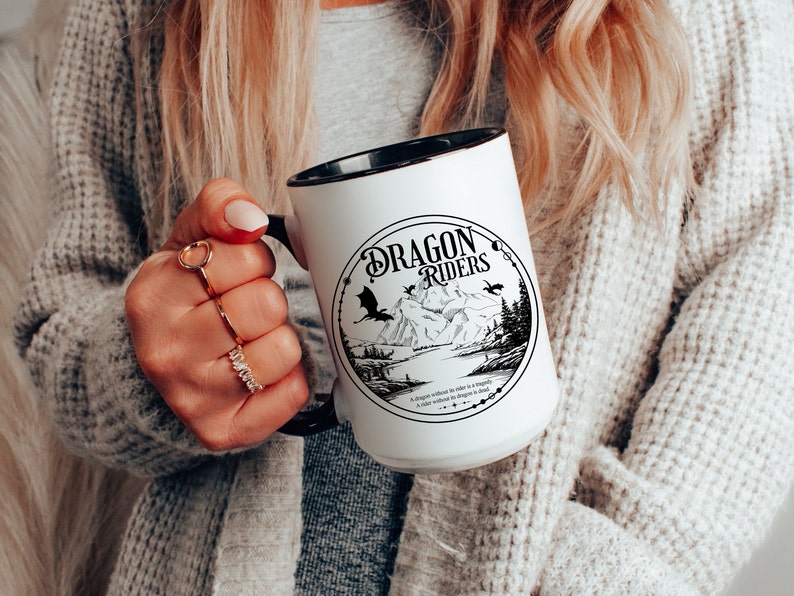 May include: White ceramic mug with a black rim and handle. The mug features a black and white illustration of a mountain scene with two dragons flying overhead. The text "Dragon Riders" is printed in a black, stylized font.
