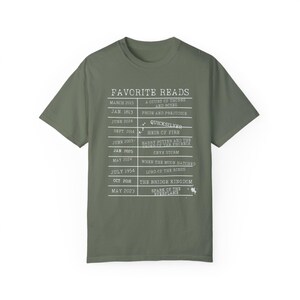 May include: A green t-shirt with a white graphic listing favorite reads and the dates they were read. The graphic includes titles like "A Court of Thorns and Roses", "Pride and Prejudice", "Quicksilver", "Heir of Fire", "Harry Potter and the Order of the Phoenix", "When the Moon Hatched", "Lord of the Rings", "The Bridge Kingdom", and "Spark of the Perflame".