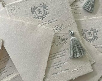 May include: A stack of white wedding invitations with a silver crest and the text "in honor of your presence at the wedding of Mary Norton and Nichols" and "OQ Farms, Bridgewater Corners, Vermont, Black Tie Optional".