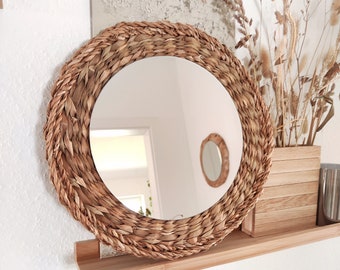 May include: A round woven mirror with a natural brown finish. The mirror is hanging on a wooden shelf with dried grasses in a vase.