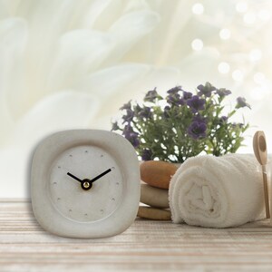 Könnte beinhalten: Eine weiße quadratische Uhr mit schwarzen Zeigern und einem goldenen Zentrum. Die Uhr steht auf einem Holztisch mit einem weißen Handtuch und einer Topfpflanze mit lila Blüten.