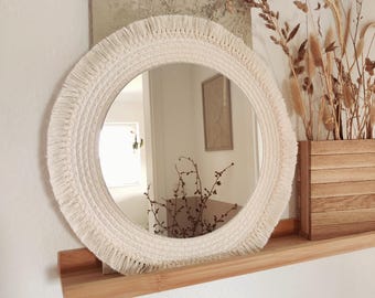 May include: A round mirror with a white macrame frame and fringe. The mirror is hanging on a wooden shelf with dried flowers in a wooden box.
