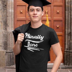May include: A person wearing a black graduation cap and gown, holding a black t-shirt with the text "Phinally Done" in white.