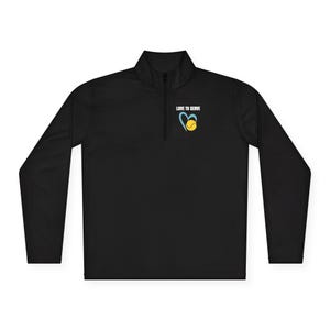 Könnte beinhalten: Schwarzes Langarm-Sportshirt mit einem Kragen mit Viertelreißverschluss. Das Shirt hat eine Grafik eines blauen Herzens mit einem gelben Tennisball darin und den Text "LOVE TO SERVE" darüber.