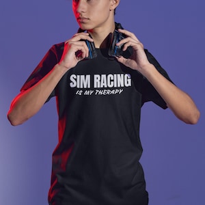 May include: A person wearing a black t-shirt with the text "Sim Racing is my therapy" in white. The person is holding a pair of black headphones with blue accents around their neck.