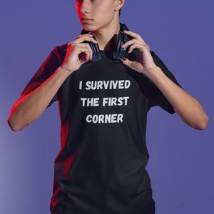 May include: A young person wearing a black t-shirt with the text "I survived the first corner" in white. They are holding a pair of black headphones with blue accents around their neck.