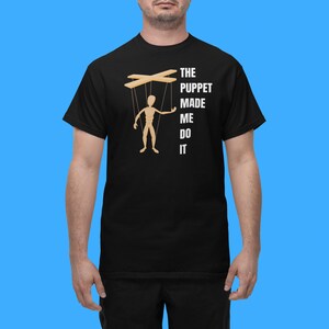 May include: A black t-shirt with a white graphic of a puppet with strings attached to its limbs. The text "THE PUPPET MADE ME DO IT" is printed below the puppet.