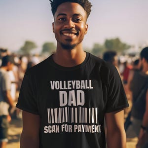 以下が含まれることがあります： 「Volleyball Dad」と白文字で書かれた黒いTシャツ。その下に「Scan for Payment」と書かれたバーコードがあります。