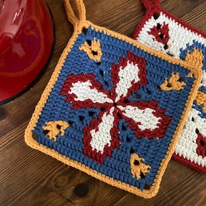 Puede incluir: Dos agarraderas de olla de ganchillo con un diseño floral. Una es azul, blanca y roja con un borde amarillo. La otra es blanca, roja y amarilla con un borde azul.