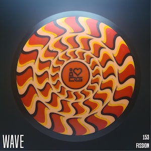 May include: A round disc golf disc with a vibrant, swirling pattern in shades of orange, yellow, and black. The center features a circular design with a heart symbol and text. The word "WAVE" is printed at the bottom left, and "153 FISSION" is at the bottom right.