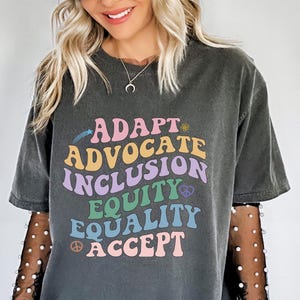 May include: A dark gray t-shirt with a graphic print of the words "Adapt", "Advocate", "Inclusion", "Equity", "Equality", and "Accept" in a rainbow of colors. The words are arranged in a curved shape.