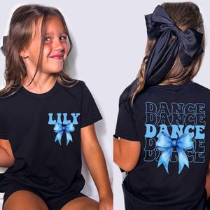 May include: Black t-shirt with a blue bow and the name "LILY" on the front. The back of the shirt features the word "DANCE" repeated four times, along with a blue bow graphic. The shirt is designed for dance.