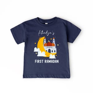 A navy blue t-shirt with a graphic design depicting a mosque under a crescent moon and stars, along with the text "Khadja's FIRST RAMADAN" printed on it.