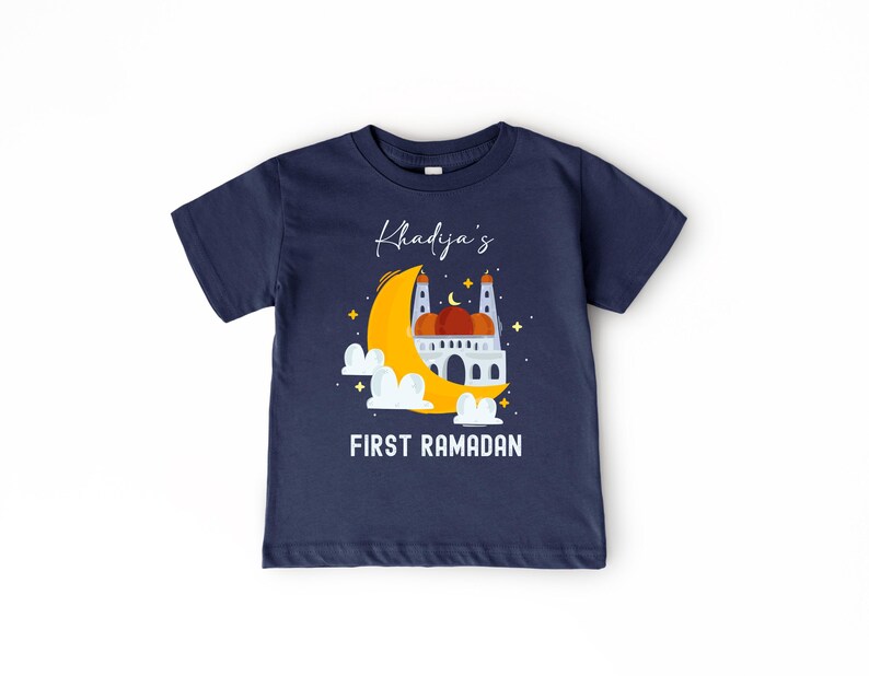 A navy blue t-shirt with a graphic design depicting a mosque under a crescent moon and stars, along with the text "Khadja's FIRST RAMADAN" printed on it.