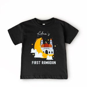 A black t-shirt with a graphic design depicting a mosque and a crescent moon, along with the text "Adam's FIRST RAMADAN" printed on it.