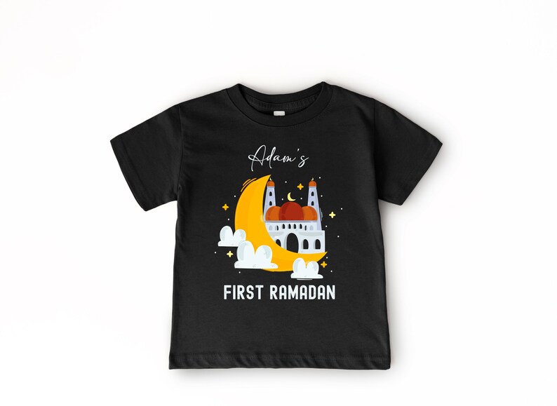 A black t-shirt with a graphic design depicting a mosque and a crescent moon, along with the text "Adam's FIRST RAMADAN" printed on it.