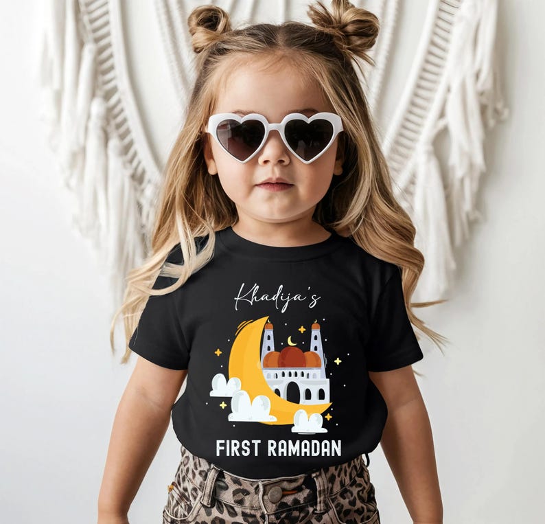 A young girl wearing a black t-shirt with a graphic design and heart-shaped sunglasses, standing in front of a white background.