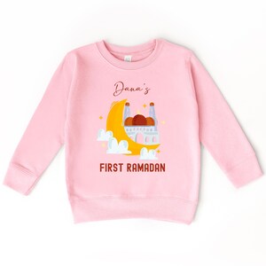 A pink long-sleeved sweatshirt with a graphic design depicting a mosque and a crescent moon, along with the text "Dana's FIRST RAMADAN".