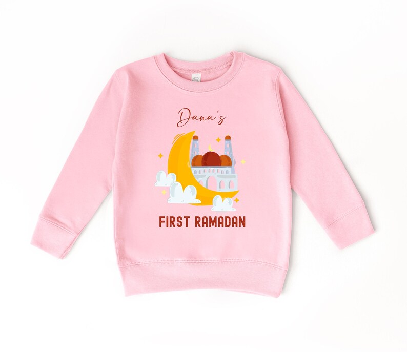 A pink long-sleeved sweatshirt with a graphic design depicting a mosque and a crescent moon, along with the text "Dana's FIRST RAMADAN".