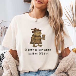 May include: Cream-colored t-shirt featuring a whimsical illustration of a frog in medieval attire, holding a goblet. The text "I have to say weird stuff or I'll die" is printed below the graphic. The shirt has a relaxed fit.