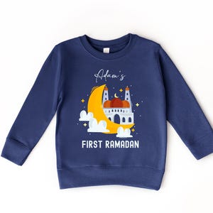A navy blue sweatshirt with a graphic design depicting a mosque and a crescent moon, along with the text "Adam's FIRST RAMADAN" printed on it.