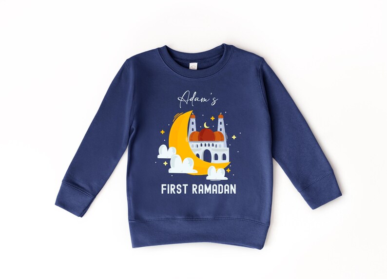 A navy blue sweatshirt with a graphic design depicting a mosque and a crescent moon, along with the text "Adam's FIRST RAMADAN" printed on it.