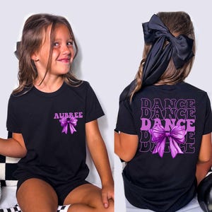 May include: Black t-shirt with a purple bow graphic and the name "AUBREE" on the front. The back of the shirt features the word "DANCE" stacked four times with a purple bow graphic. The girl is wearing a black bow in her hair.