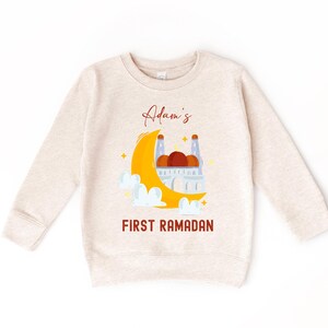 A beige sweatshirt with a graphic design depicting a crescent moon, a mosque, and clouds, along with the text "Adam's FIRST RAMADAN" printed on it.