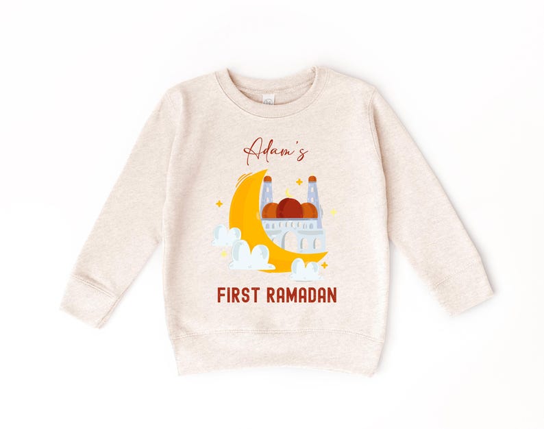 A beige sweatshirt with a graphic design depicting a crescent moon, a mosque, and clouds, along with the text "Adam's FIRST RAMADAN" printed on it.