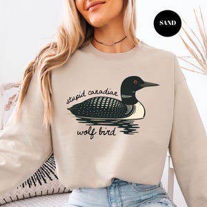 May include: Sand-colored sweatshirt featuring a black and white illustration of a loon on water, with the text "stupid canadian wolf bird" in black script. The loon has a black head, back, and beak, with a white chest and a red eye.
