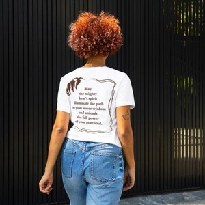 May include: White t-shirt with a brown graphic print of a bear and text that reads "May the mighty bear's spirit illuminate the path to your inner wisdom and unleash the full power of your potential."