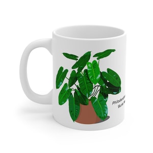 Peut inclure: Une tasse en céramique blanche avec une illustration d'une plante verte Philodendron 'Birkin'. La plante est dans un pot brun et le texte 'Philodendron 'Birkin'' est imprimé sous la plante.