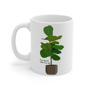 Könnte beinhalten: Weiße Keramiktasse mit einer Illustration einer grünen Geigenfeige. Der Text "Ficus lyrata 'fiddle leaf fig'" ist unter der Pflanze aufgedruckt.