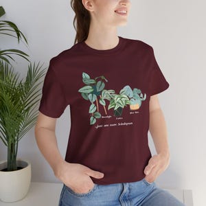 以下が含まれることがあります： 3つのScindapsus植物の植物図が描かれた、マルーン色のTシャツ。「...just one more Scindapsus」というテキストが入っています。植物には「Moonlight」、「Exotica」、「Silver Hero」とラベルが付けられています。