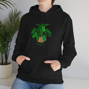 Peut inclure: Sweat à capuche noir avec une illustration de plante verte dans un pot marron. Le texte "Philodendron Lobe Burke Mare" est imprimé en dessous. Le sweat-shirt a une poche avant et une capuche à cordon.