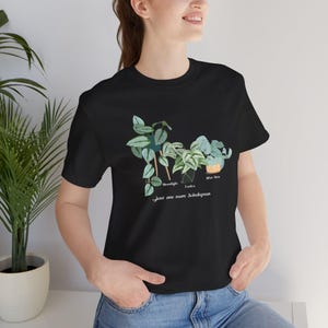 以下が含まれることがあります： 植物のグラフィックがプリントされた黒いTシャツ。植物は3種類あり、それぞれ「Monstera Deliciosa」、「L'unica」、「Silver Satin」とラベル付けされています。植物の下には「...just one more Scindapsus」というテキストがプリントされています。