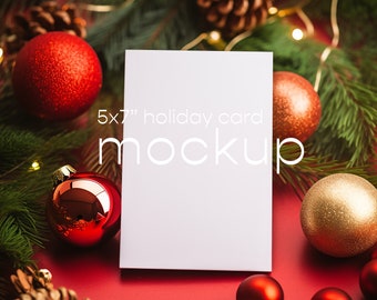 May include: A white 5x7 inch holiday card mockup surrounded by red and gold ornaments and pine branches with twinkling lights.