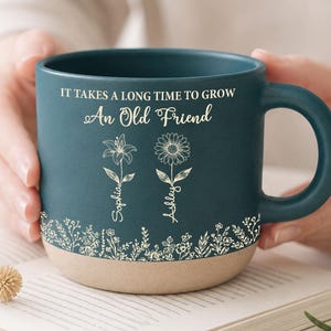 May include: Teal ceramic mug with a beige base, featuring the text "IT TAKES A LONG TIME TO GROW An Old Friend." Floral illustrations and names "Sophia" and "Ashley" are also present. The mug is held over an open book.