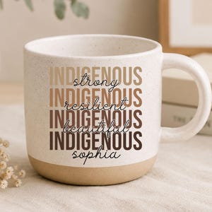 May include: A speckled white ceramic mug with a tan base and a curved handle. The mug features the word "INDIGENOUS" repeated in a stacked design, with words like "strong" and "beautiful" in between. The name "Sophia" is at the bottom.