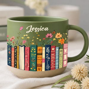 May include: A green ceramic mug with a floral and book design. The mug features a row of colorful books at the base and a field of flowers. The name "Jessica" is written in script on the front.