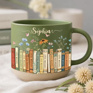 May include: A green ceramic mug with a beige base, featuring a floral and book design. The mug has the name "Sophia" in script and the words "Custom Text" on the books. The mug has a large handle and is decorated with flowers and butterflies.