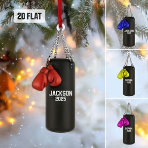 May include: A black punching bag ornament with red boxing gloves, hanging from a silver chain and red ribbon. The ornament has the name "JACKSON" and the year "2025" printed in white. The background is a snowy evergreen branch.