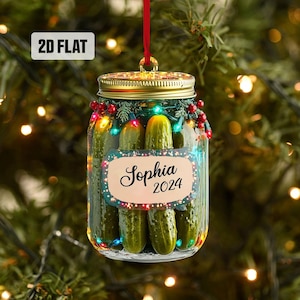 May include: A 2D flat Christmas ornament shaped like a jar of pickles. The jar has a gold lid and is filled with green pickles. The ornament is personalized with the name "Sophia" and the year "2024". It is decorated with string lights and red berries.