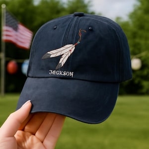 May include: Navy blue baseball cap with an embroidered feather design and the name "JACKSON" in white. The feathers are white with black tips and red and brown accents. The cap is held up by a hand.