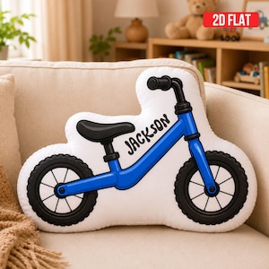 May include: A white throw pillow shaped like a balance bike with a blue frame, black seat, and black tires. The name Jackson is printed in black script on the frame. The pillow features a 2D flat design with clear black outlines.